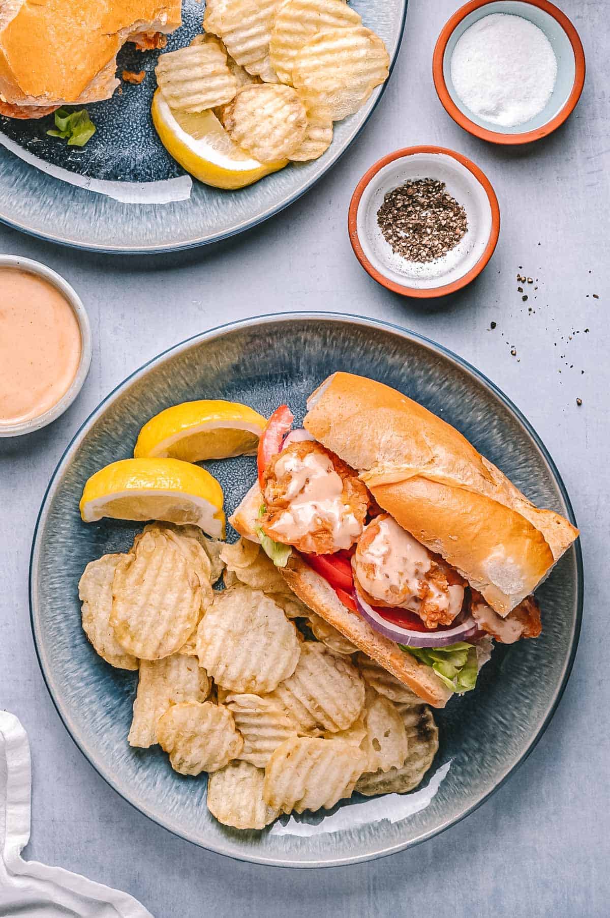 Overhead shot of a shrimp po'boy sandwich with ridged potato chips and lemon wedges on a blue plate, with bowls of salt, pepper, and dipping sauce in the background.