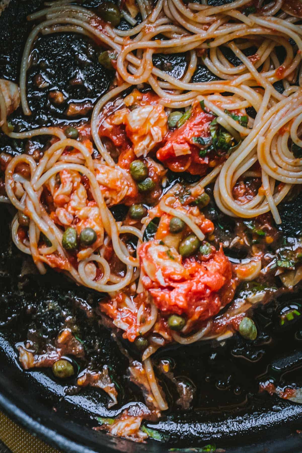 Spaghetti glistens in a deep, rustic skillet, coated in a rich tomato sauce with visible chunks of fish and plump capers. The pan's dark surface highlights the vibrant reds and greens of the dish's fresh ingredients.