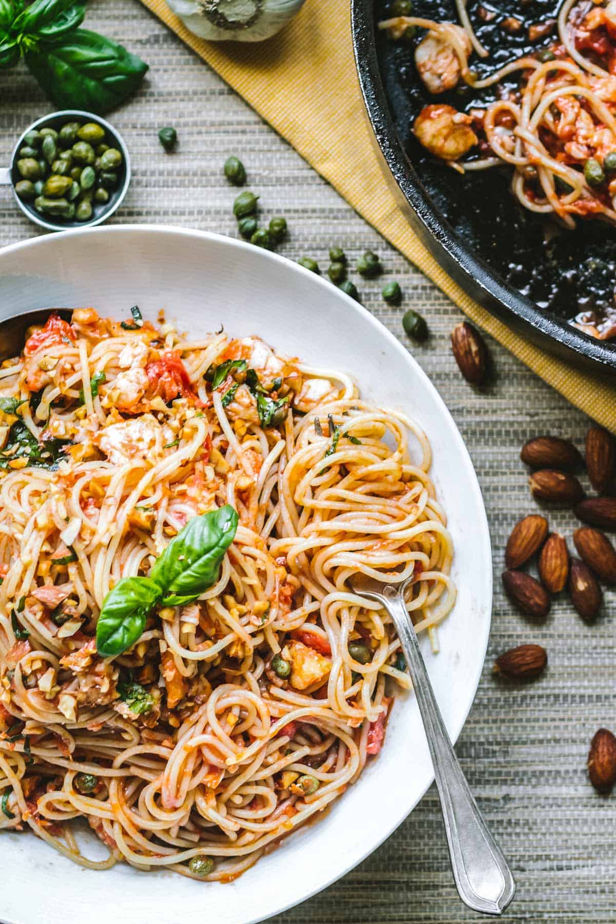 A white bowl brimming with saucy spaghetti sits at the center of a cozy tabletop. Surrounding it are raw almonds, capers, and garlic cloves-hinting at the Mediterranean ingredients used in the dish.