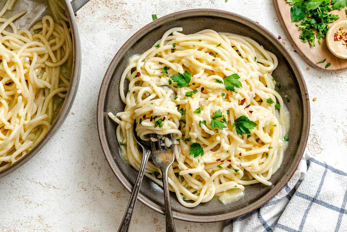 Overhead view of a table setting with a bowl of pasta aglio e olio in the foreground and a larger serving pot visible in the background. The elegant yet simple presentation shows the pasta garnished with parsley and red pepper flakes, with utensils ready for dining.