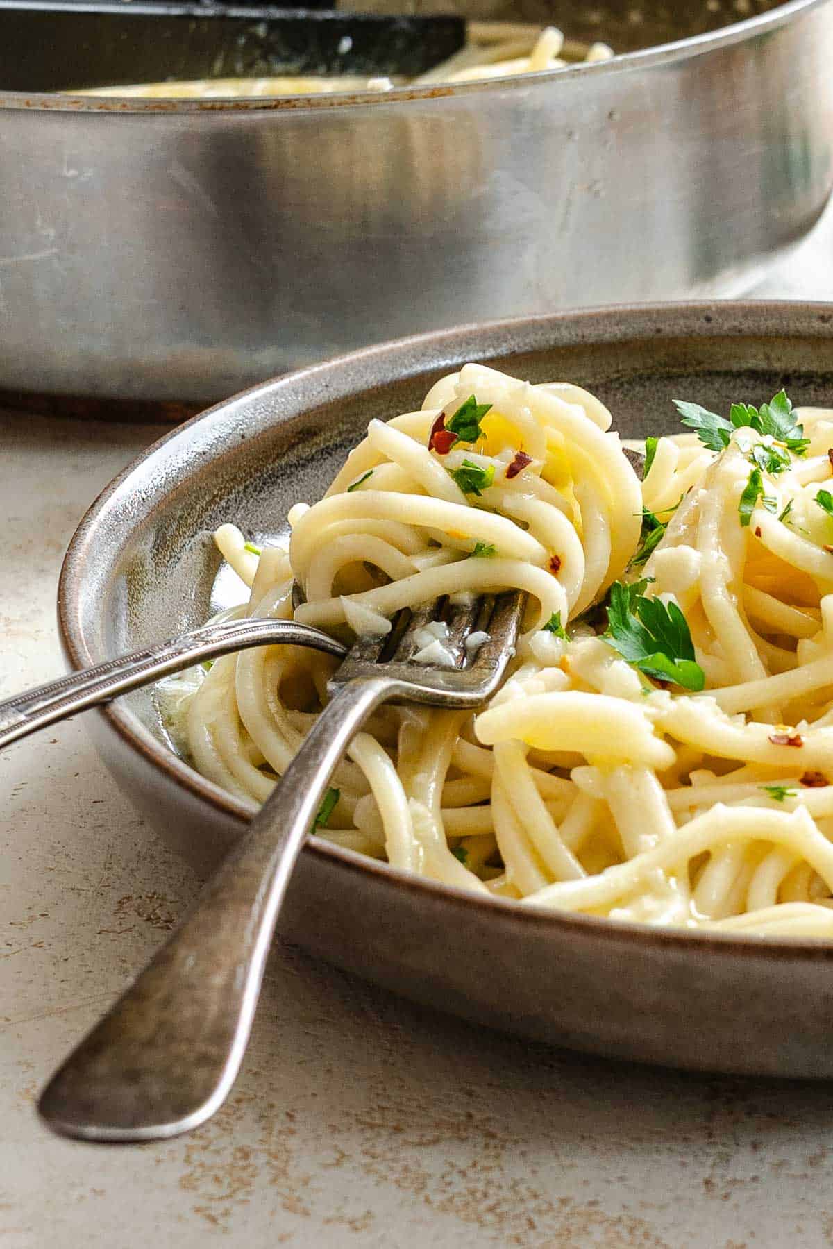 An artistic angled close-up of pasta aglio e olio being twirled on a fork, showcasing the silky texture of the pasta coated in flavorful olive oil. The shallow depth of field emphasizes the fork lifting the pasta from the ceramic dish, with the cooking pot blurred in the background.