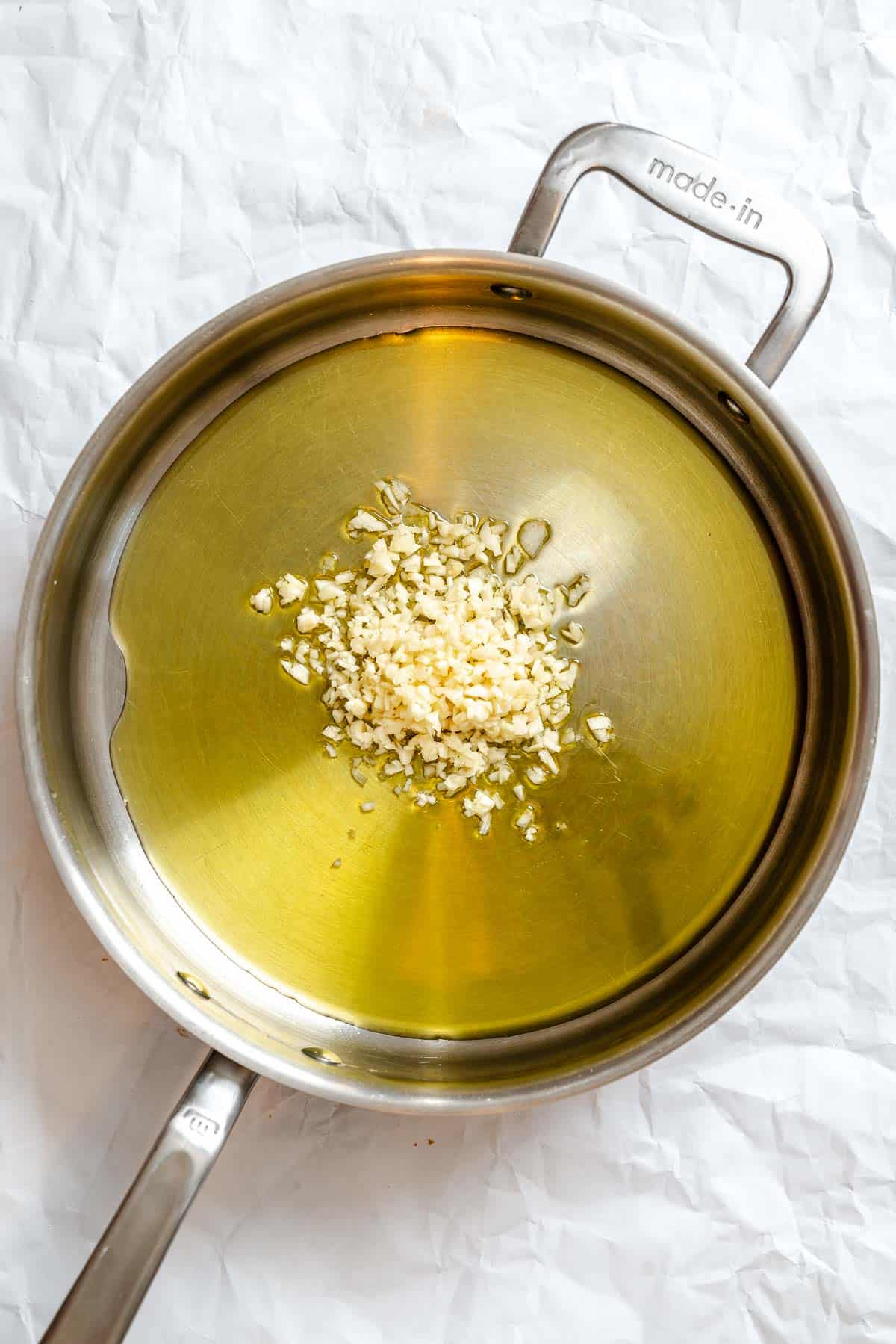 Overhead view of a stainless steel saucepan containing minced garlic beginning to cook in olive oil. The garlic appears freshly added and not yet sautéed, capturing the first cooking stage of the dish where the aromatics infuse the oil.