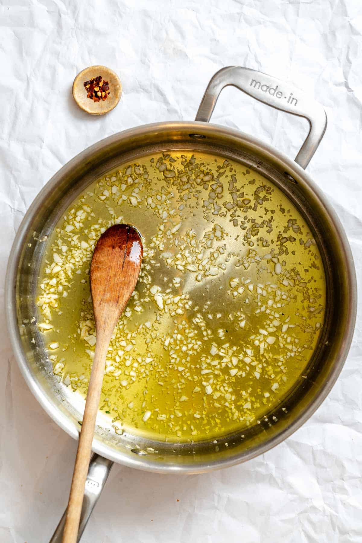 A saucepan with golden minced garlic sautéed in olive oil, with red pepper flakes visible throughout the shimmering oil. A wooden spoon rests in the pan, showing how the garlic has been sautéed to a perfect light golden color without burning.