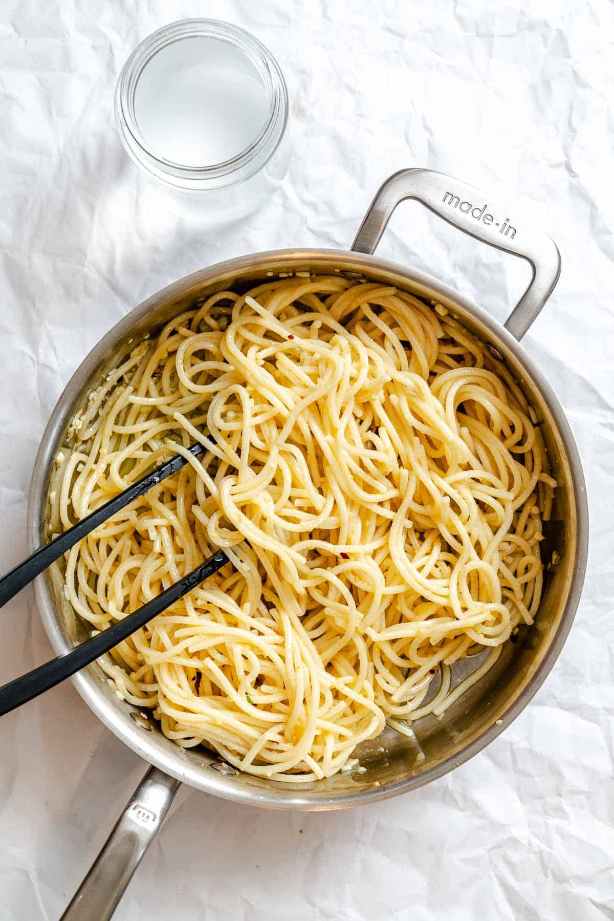 Overhead view of freshly cooked spaghetti in a stainless steel pot, tossed with tongs to show the pasta's perfectly cooked texture. The image demonstrates how the pasta should look before adding it to the garlic-infused oil.