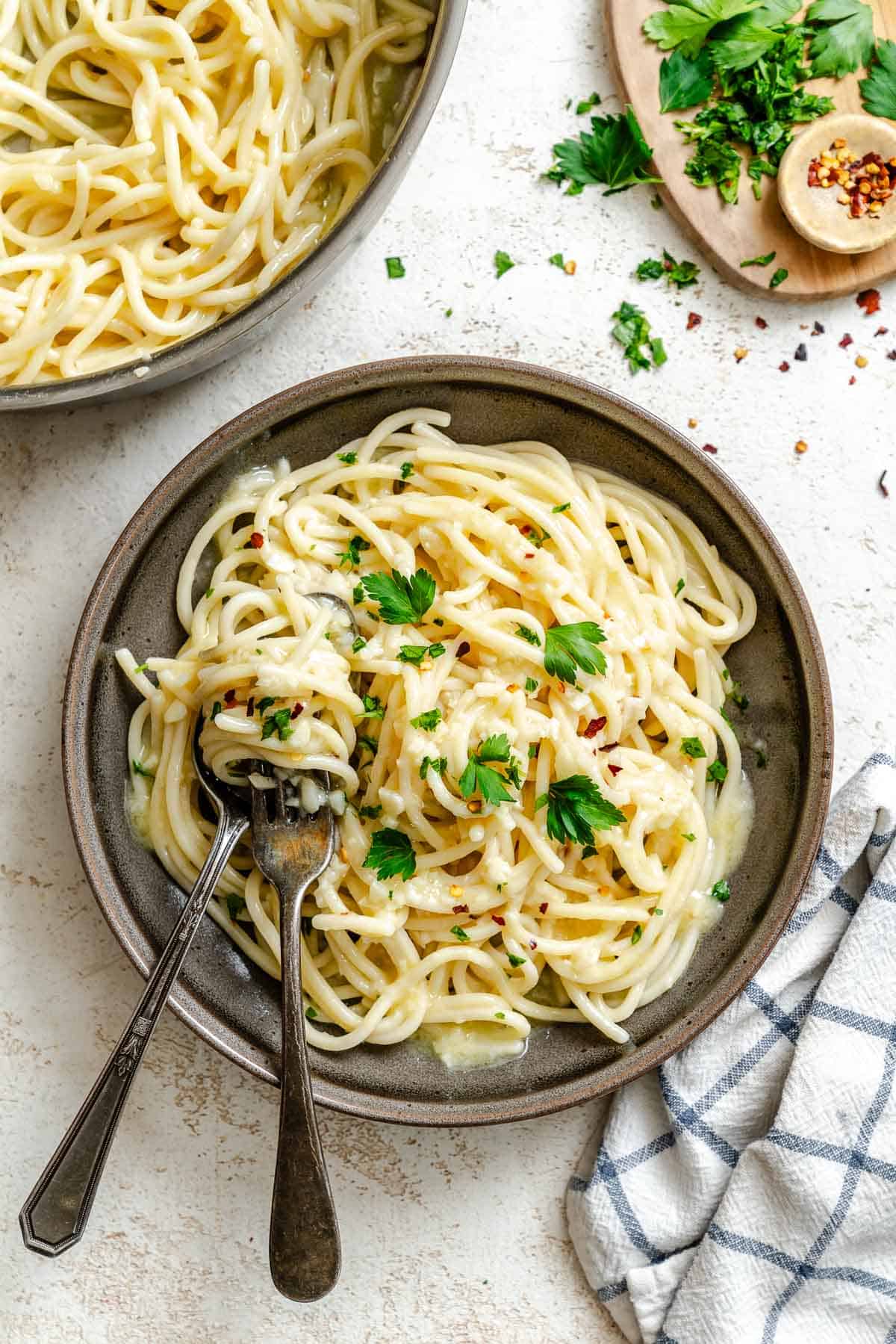 The finished pasta aglio e olio served in a rustic ceramic bowl and garnished with fresh parsley. The glossy spaghetti shows traces of the garlic, oil, and red pepper flakes coating each strand. A pair of serving utensils rest in the dish, ready for serving.
