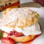 Close-up of a split shortcake biscuit topped with whipped cream and fresh strawberries, dusted with powdered sugar. A bottle of whiskey is partially visible in the background, suggesting a boozy addition to the dessert.
