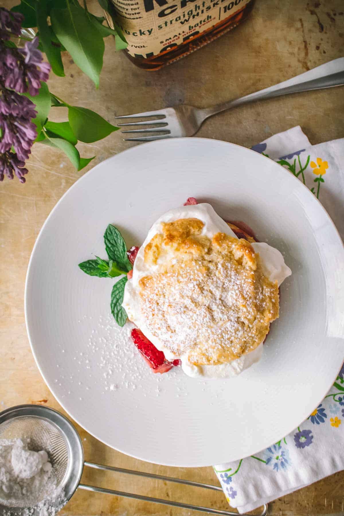 Overhead view of a strawberry shortcake on a white plate, garnished with fresh mint leaves. The golden biscuit is dusted with powdered sugar and strawberry juices peek out from beneath. A bourbon bottle, purple flowers, and kitchen tools frame the composition.