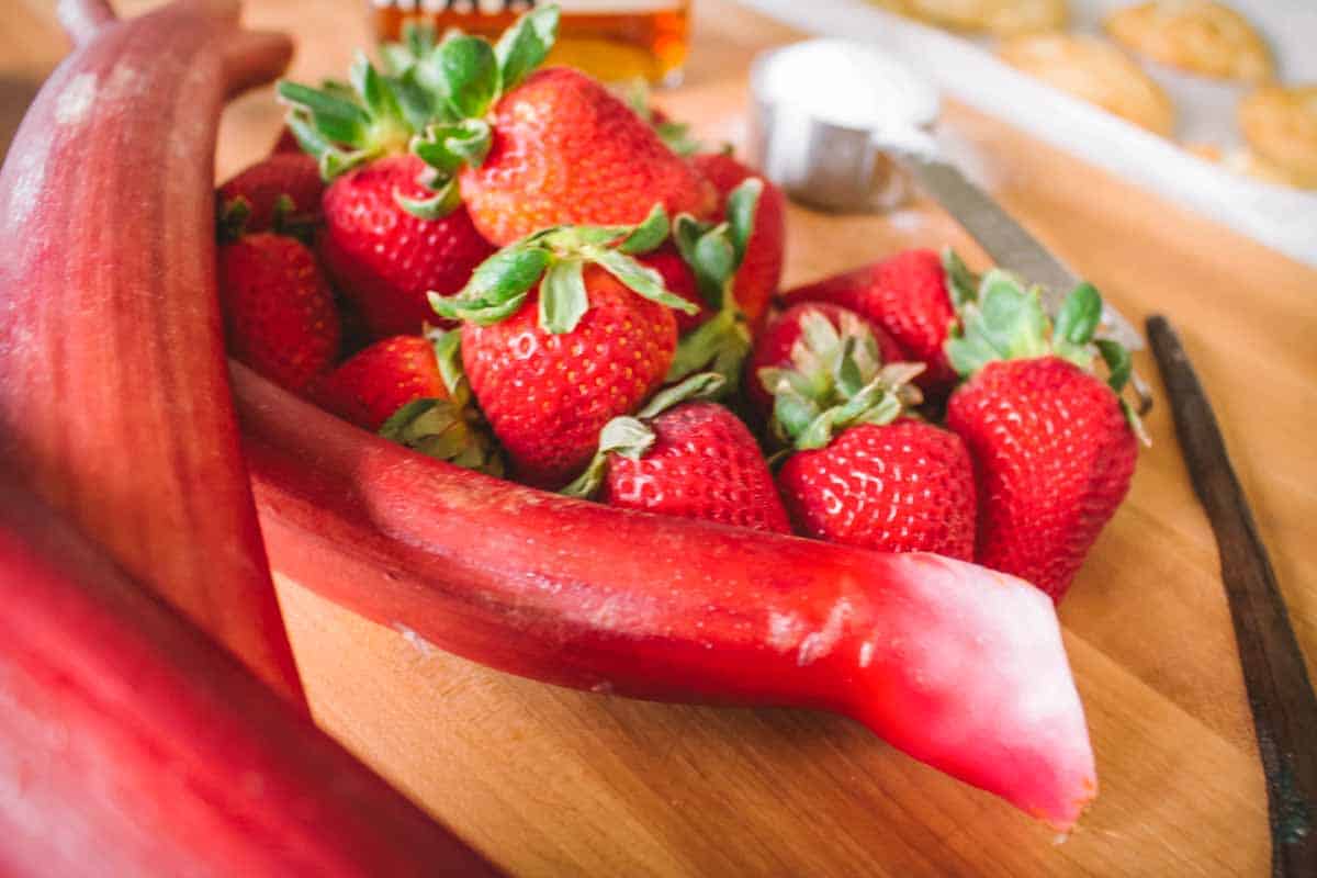 Fresh ingredients for the recipe - vibrant red strawberries nestled with slices of bright pink rhubarb on a wooden cutting board. The rhubarb's distinctive color contrasts with the berries, with baking supplies visible in the background.
