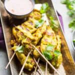 Grilled chicken satay skewers arranged on a hammered copper tray, garnished with fresh cilantro and served with a small bowl of creamy peanut dipping sauce.