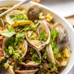 Clams in a white bowl with charred corn, chorizo, and herbs, served with rustic bread.