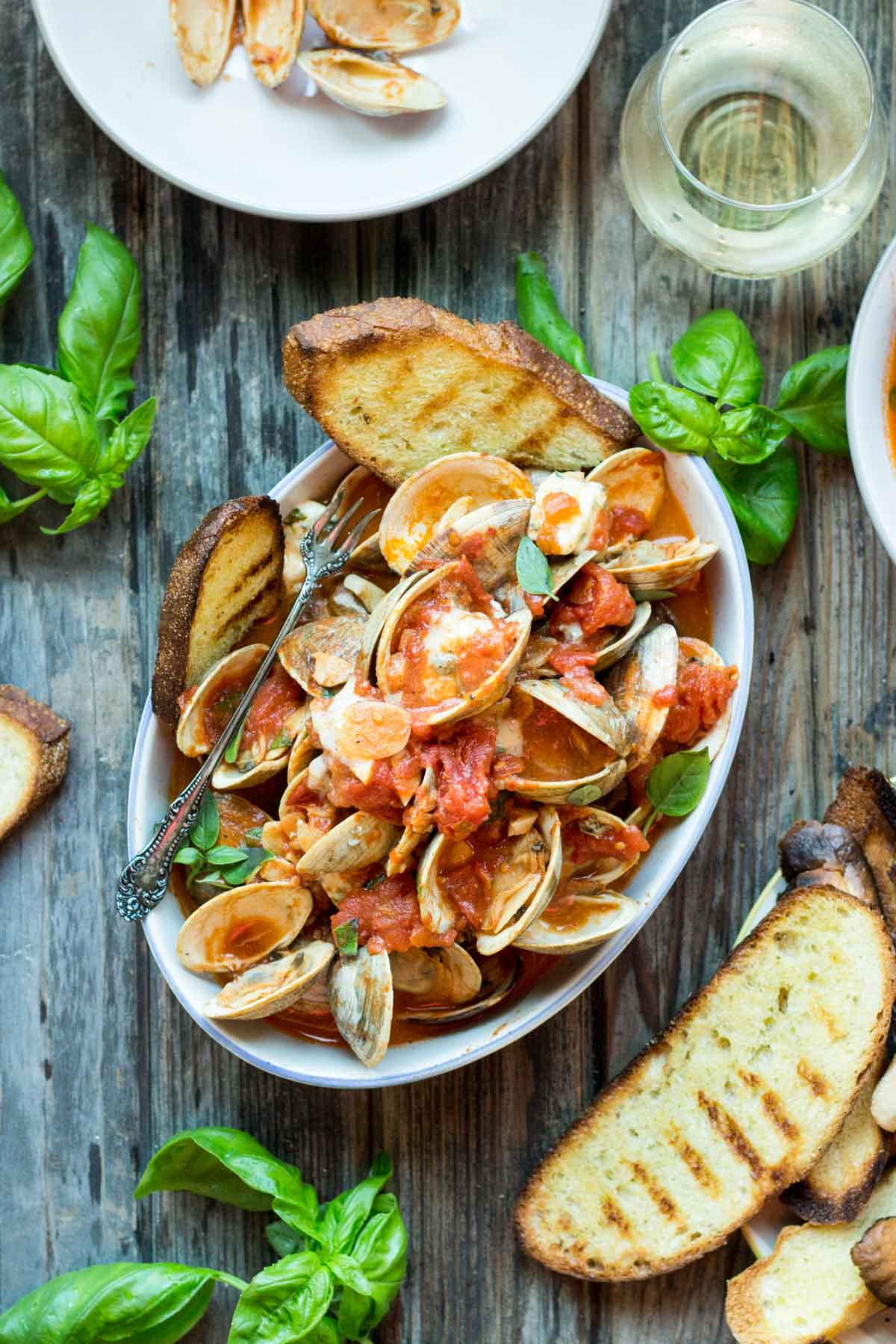 Overhead view of clams in tomato sauce with fresh basil leaves and blue cheese crumbles, served with toasted bread slices in a white bowl on a rustic wooden surface.