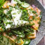 Top-down view of a vibrant salad made with sliced cantaloupe, cucumbers, and onions, topped with two stacked wedges of feta and a generous sprinkle of herbs in a decorative ceramic bowl.
