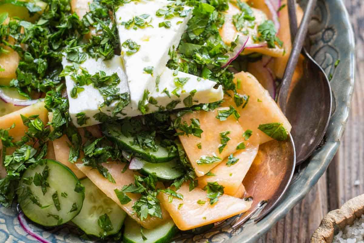 Close-up of a cucumber melon salad with cantaloupe slices, cucumber rounds, red onion, and creamy feta, generously topped with finely chopped fresh herbs.