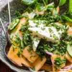 Overhead view of a colorful cucumber melon salad in a patterned ceramic bowl, garnished with fresh basil and a large wedge of feta, surrounded by sliced cucumbers and red onions.