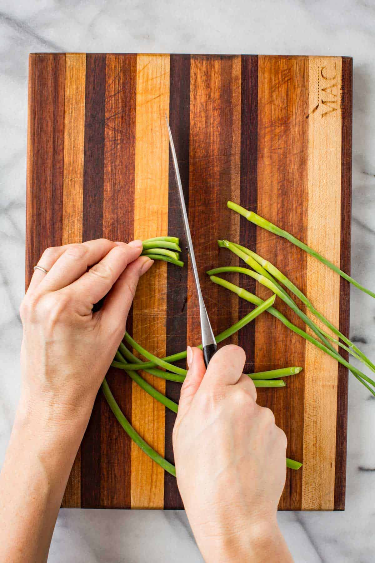 Close-up of hands using a sharp knife to cut fresh garlic scapes into small pieces on a two-tone wood cutting board.