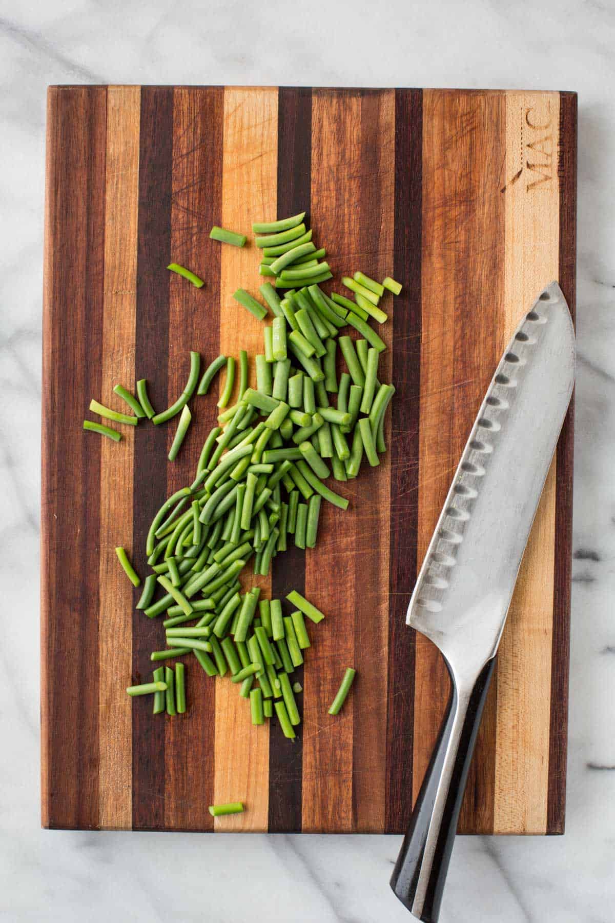 Top-down view of a pile of chopped garlic scapes next to a large chef's knife on a striped cutting board.