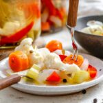 Close-up of a fork lifting a slice of pickled carrot from a plate of colorful giardiniera - includes cauliflower, red pepper, celery, and onion, lightly dressed in brine with visible herbs and peppercorns.