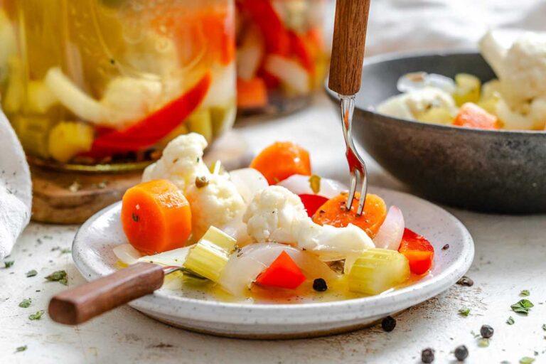Close-up of a fork lifting a slice of pickled carrot from a plate of colorful giardiniera - includes cauliflower, red pepper, celery, and onion, lightly dressed in brine with visible herbs and peppercorns.