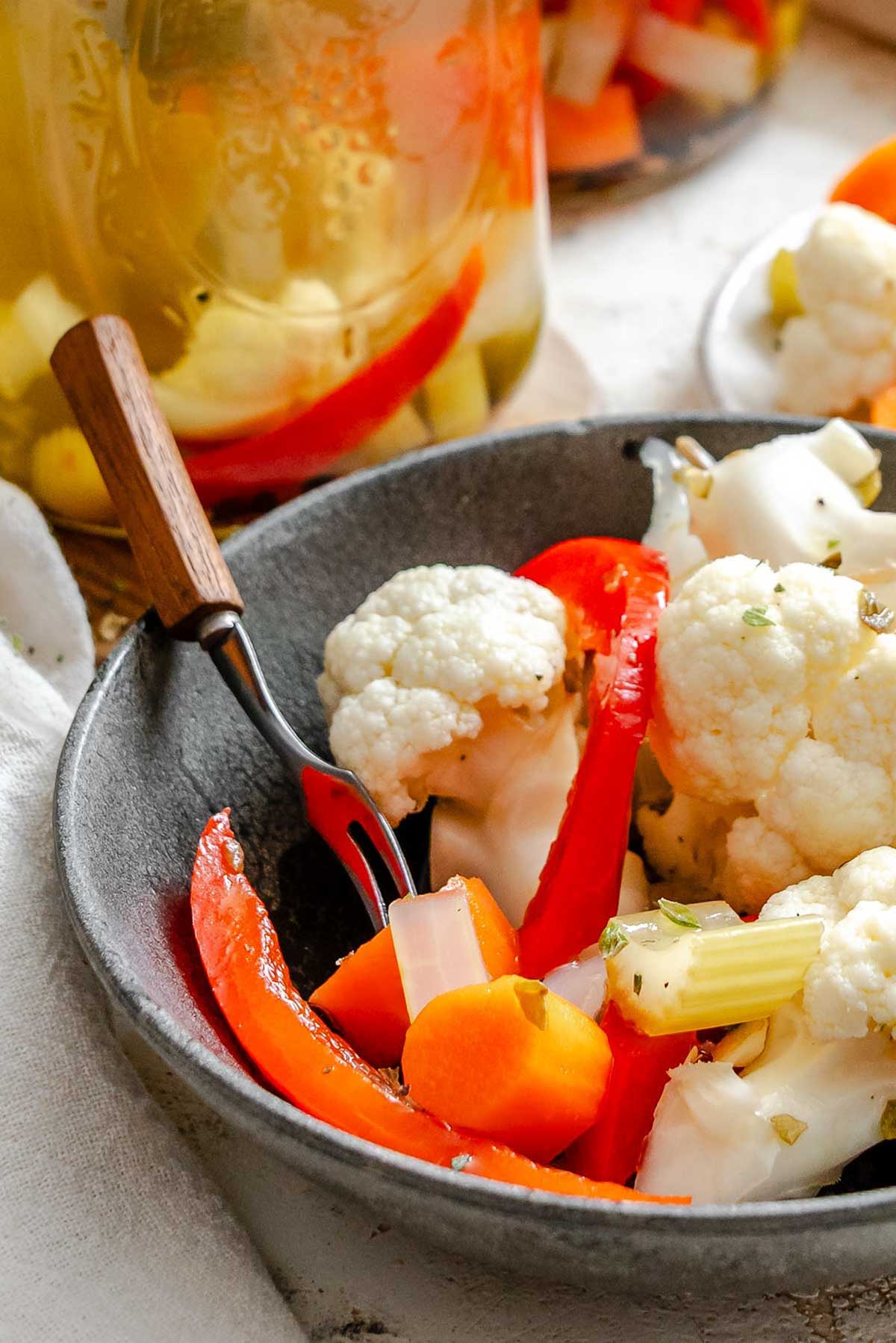 Bowl of giardiniera with fork resting inside - features vibrant chunks of pickled vegetables like cauliflower, carrots, red bell pepper, and onion in a black bowl.