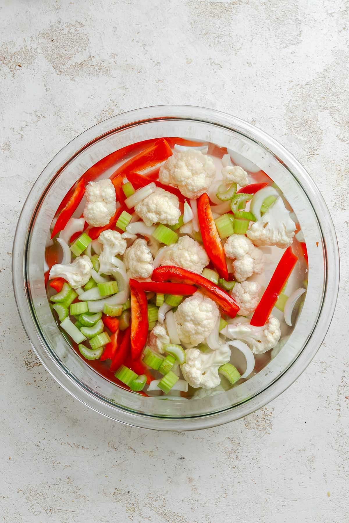 Overhead view of chopped raw vegetables in a glass bowl soaking in a brine - shows cauliflower florets, celery slices, red bell pepper strips, and onion.
