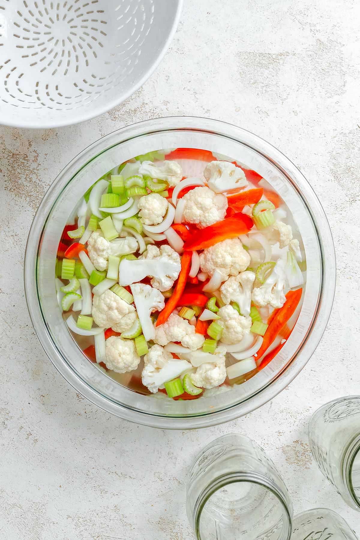 Wide shot of vegetables soaking in brine with colander and mason jars nearby - prep stage for giardiniera with visible red peppers, cauliflower, onions, and celery.