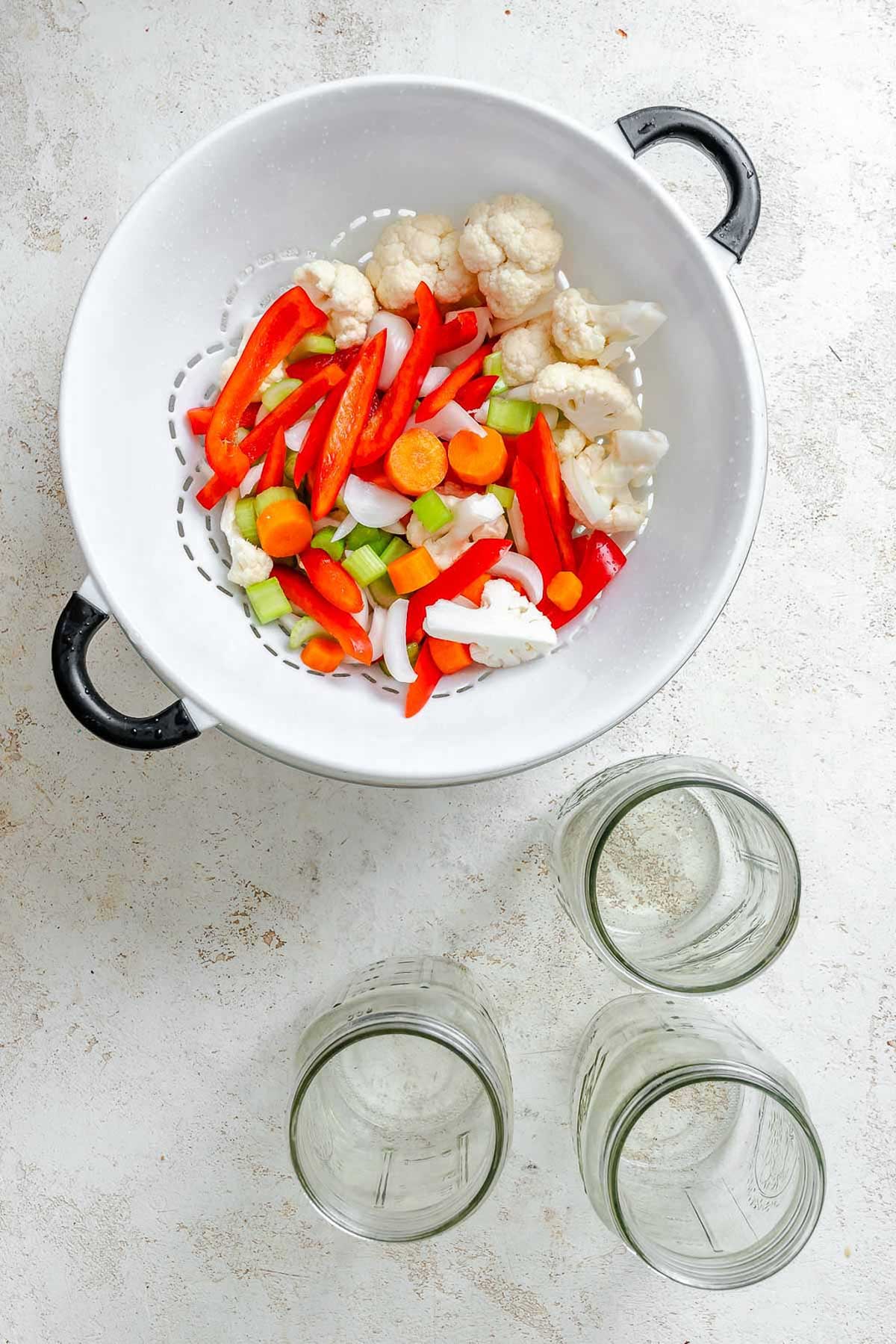 Strained giardiniera vegetables in a white colander with black handles - vibrant mix of chopped red peppers, carrots, celery, onions, and cauliflower.