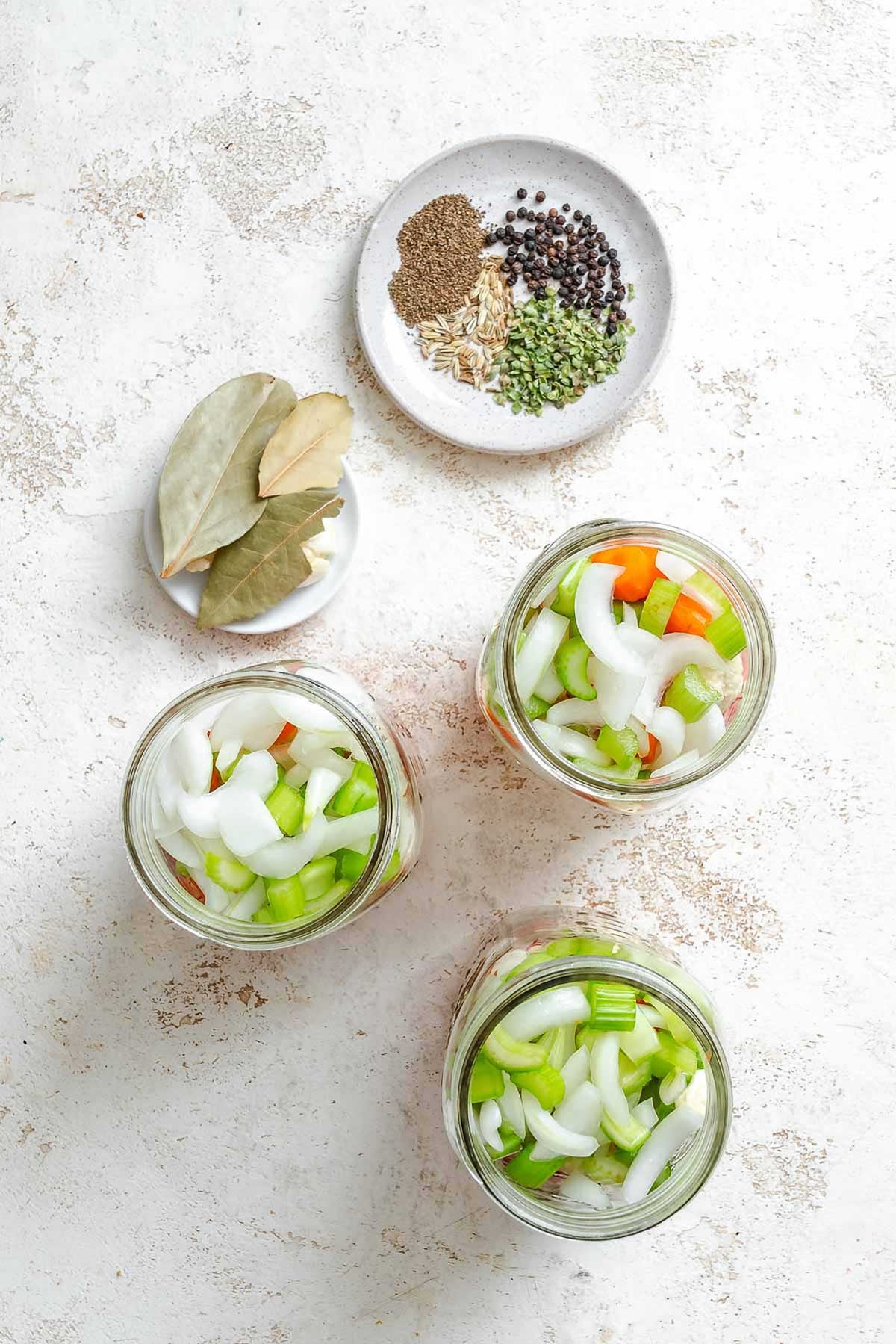 Three mason jars being filled with chopped vegetables and a plate of pickling spices nearby - visible black peppercorns, bay leaves, fennel seeds, oregano, and mustard seed.