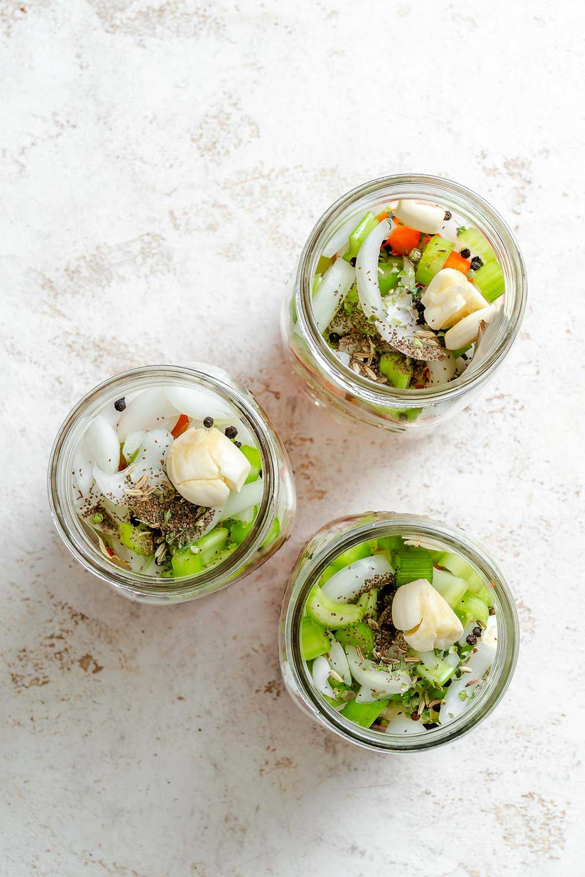 Close-up of filled jars topped with garlic cloves and seasoning - fresh giardiniera vegetables layered with whole garlic cloves, black peppercorns, and dried herbs.