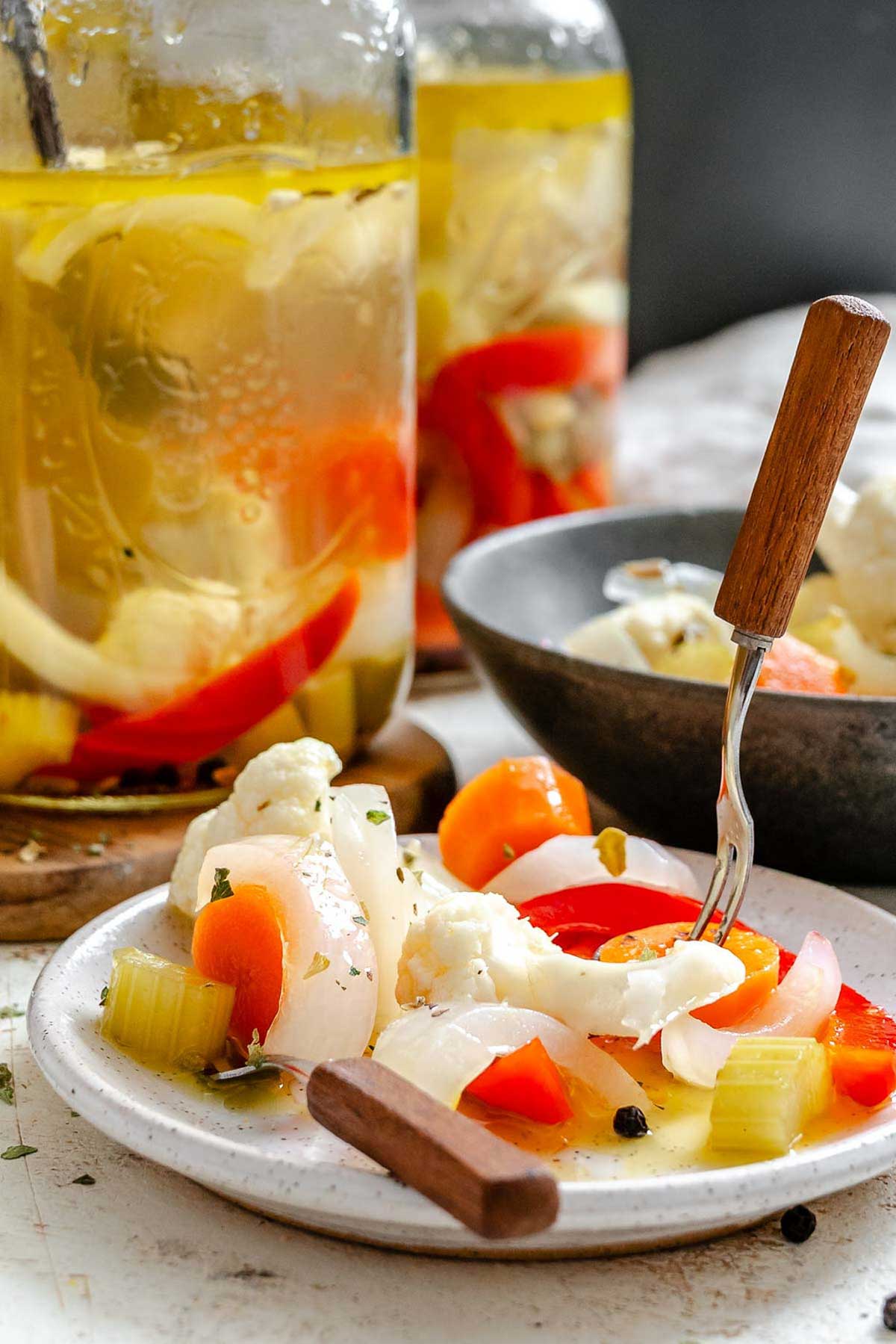 Serving plate of giardiniera with jars in background - marinated cauliflower, carrot, celery, red pepper, and onion on a white plate with wood-handled fork.