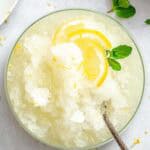 Overhead view of lemon granita in a bowl, topped with lemon slices and fresh mint, set against a bright background with scattered zest and leaves.