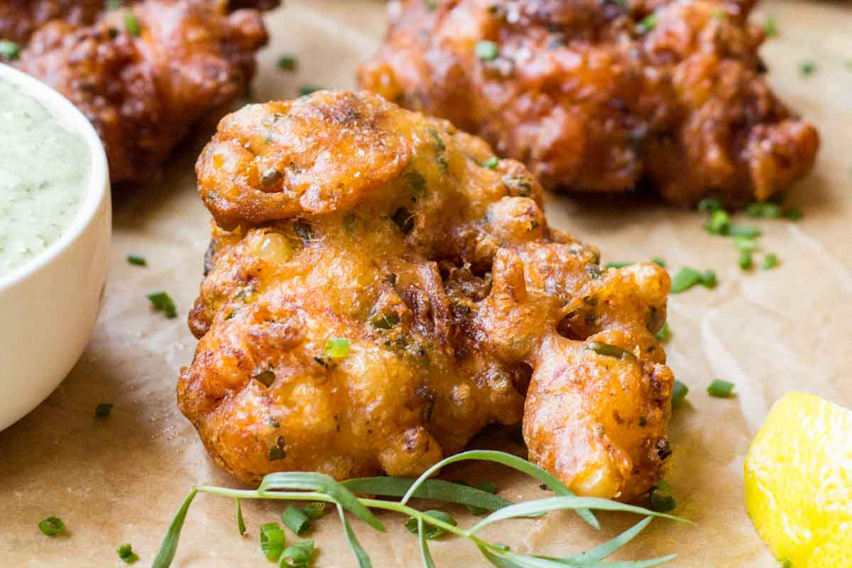 Close-up of a single crispy fritter, revealing a golden, craggy texture flecked with herbs beside a bowl of creamy aioli.