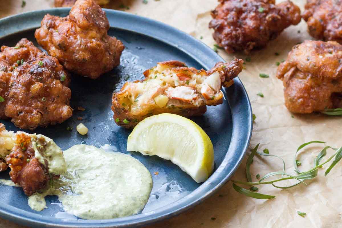 Side view of a bitten fritter on a blue plate showing juicy lobster meat and corn inside, served with a generous spoonful of aioli and a lemon wedge.