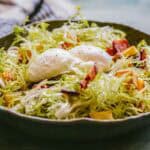 Side view of a Lyonnaise salad topped with two poached eggs nestled in a bed of frisée, with visible crispy bacon and potato croutons throughout.
