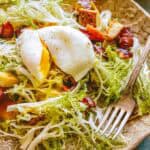 Close-up of a poached egg with its yolk beginning to ooze out over frisée lettuce, surrounded by crispy bacon and browned potato cubes.