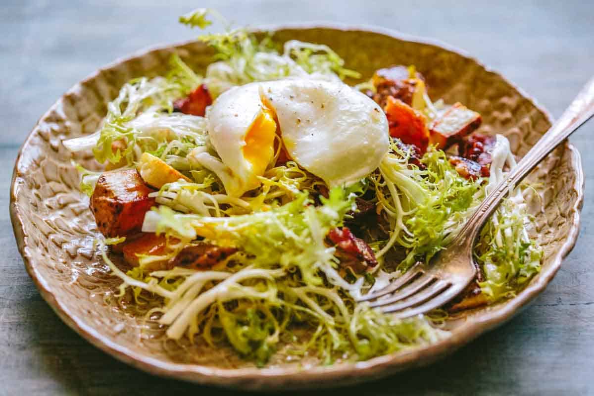 Angle view of a single poached egg split open over a rustic plate of frisée lettuce, with bits of bacon and crispy roasted potatoes scattered throughout.