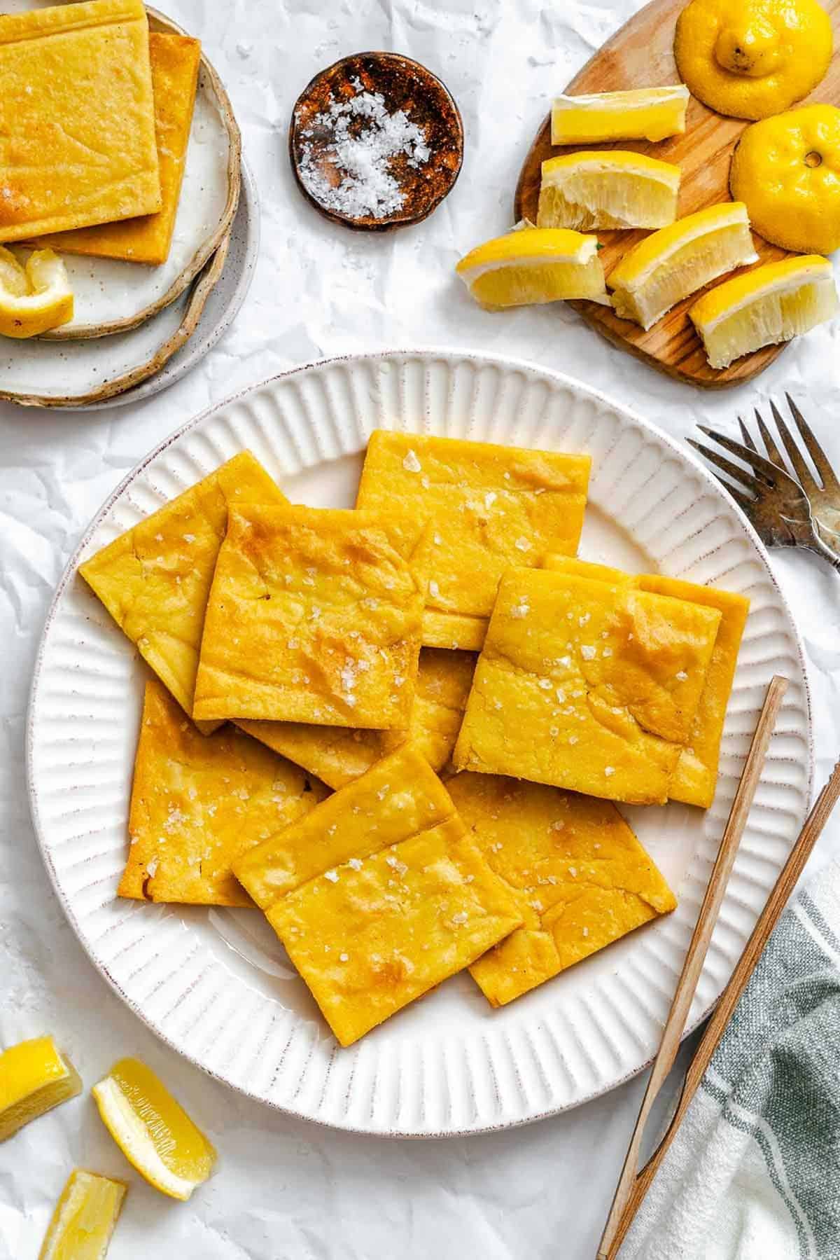 Bright overhead photo showing a plate of golden chickpea fritters surrounded by sliced lemons, salt, and cutlery-set for sharing or snacking.