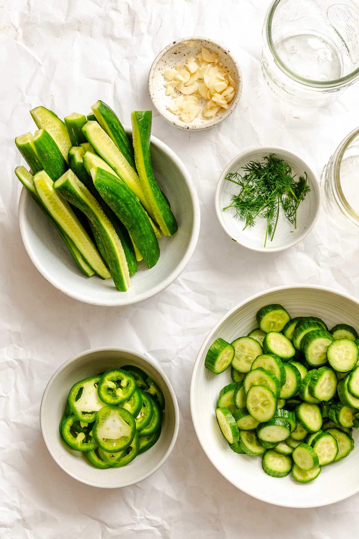 Bowls of sliced cucumbers, jalapeños, fresh dill, and garlic prepped for pickling, with empty jars nearby.