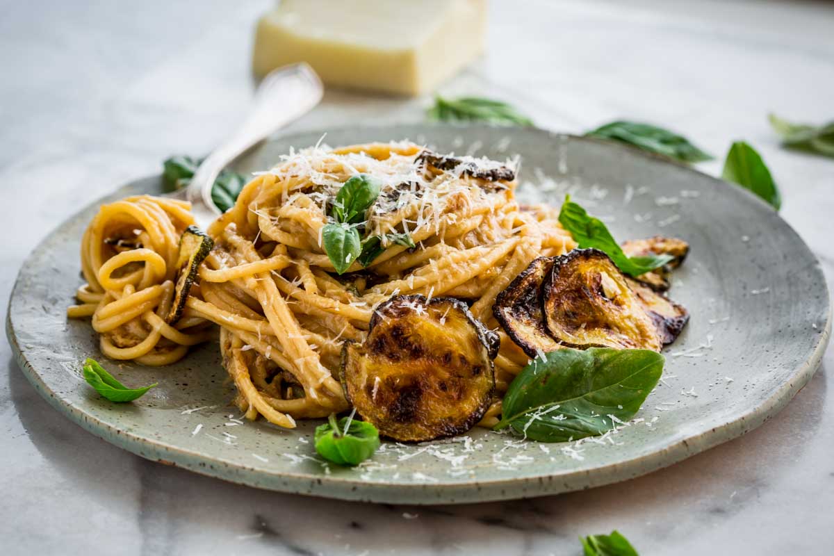A side view of spaghetti alla Nerano plated high with visible crispy zucchini rounds, fresh basil, and grated cheese, with a wedge of Pecorino Romano and scattered basil in the blurred background.