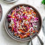 A colorful cabbage slaw with shredded red and green cabbage, carrots, and fresh herbs served in a ceramic brown bowl with silver serving utensils.