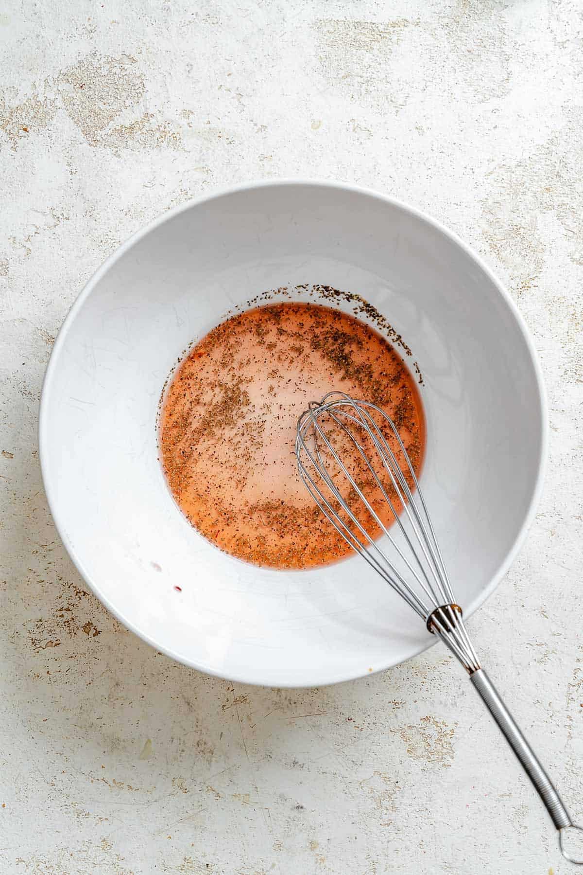 A large white mixing bowl with a reddish vinaigrette dressing speckled with black pepper, shown with a wire whisk resting inside.