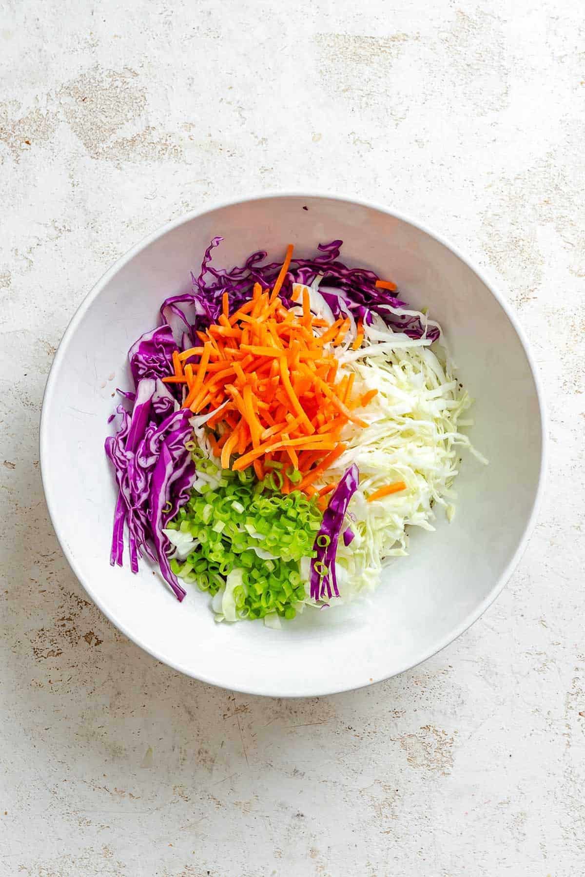 A bowl filled with neat piles of shredded red and green cabbage, julienned carrots, and sliced green onions before tossing.