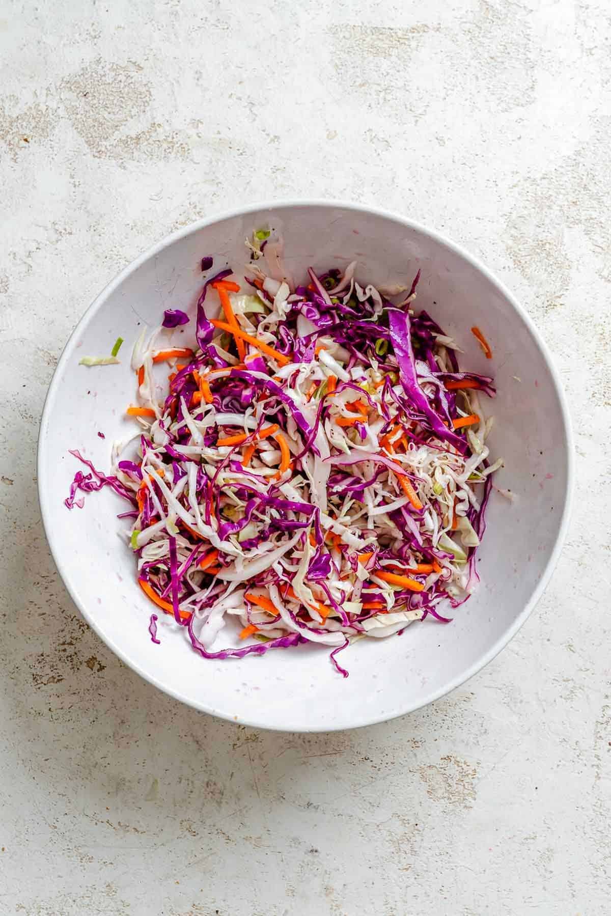 The colorful shredded cabbage and carrots are mixed together in a large white bowl, forming the slaw base.