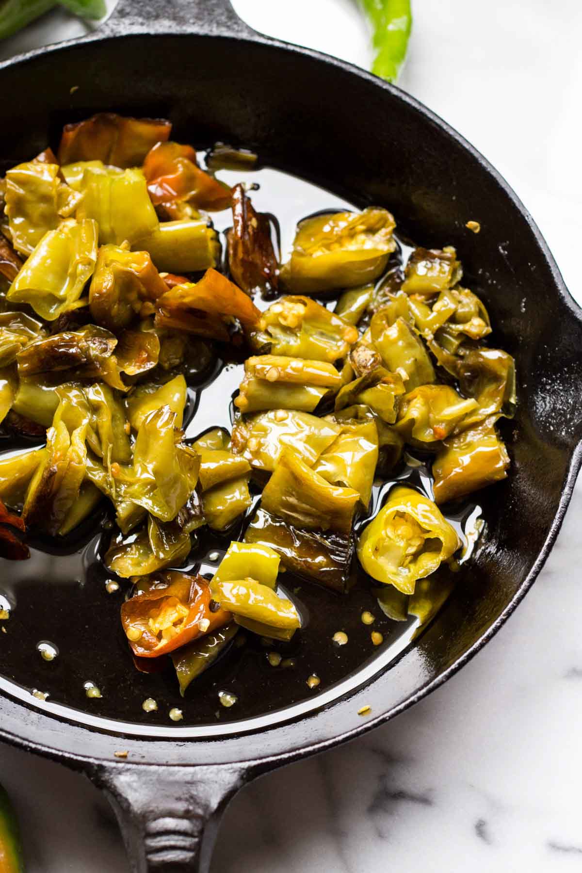 Close-up of chopped long hot peppers sizzling in oil inside a cast iron skillet, showing their blistered skins and glossy surface.
