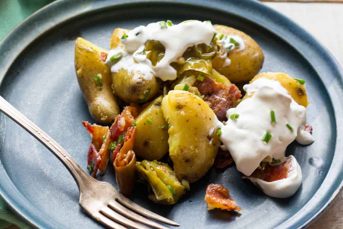 Portion of potato salad served on a dark blue plate with a fork, capturing the rustic texture of the potatoes, crispy bacon, and a smear of sour cream.