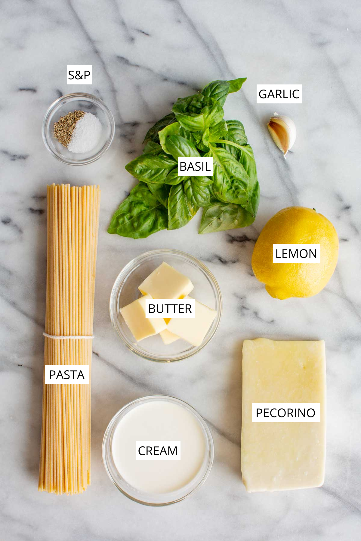 Flat lay of labeled ingredients for creamy Pasta al Limone on a marble surface, including dry spaghetti, fresh basil leaves, garlic clove, lemon, unsalted butter cubes, heavy cream, a wedge of Pecorino Romano cheese, and a bowl of salt and black pepper.