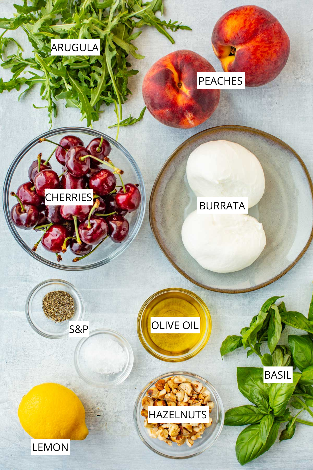 Overhead layout of labeled ingredients: arugula, peaches, cherries, burrata, olive oil, basil, lemon, hazelnuts, and salt and pepper.