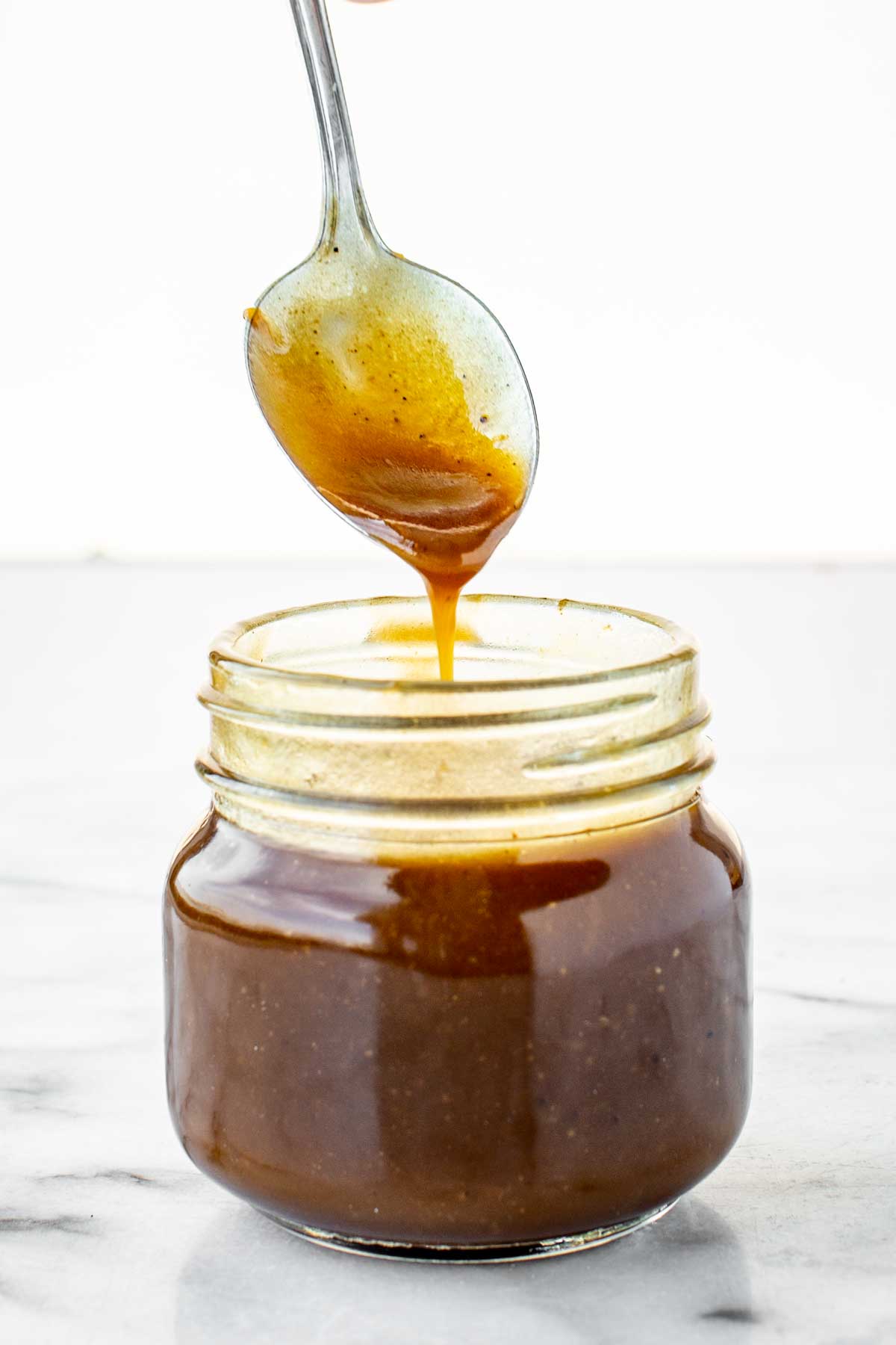 A spoon drizzling finished balsamic vinaigrette back into a glass jar with thick consistency.