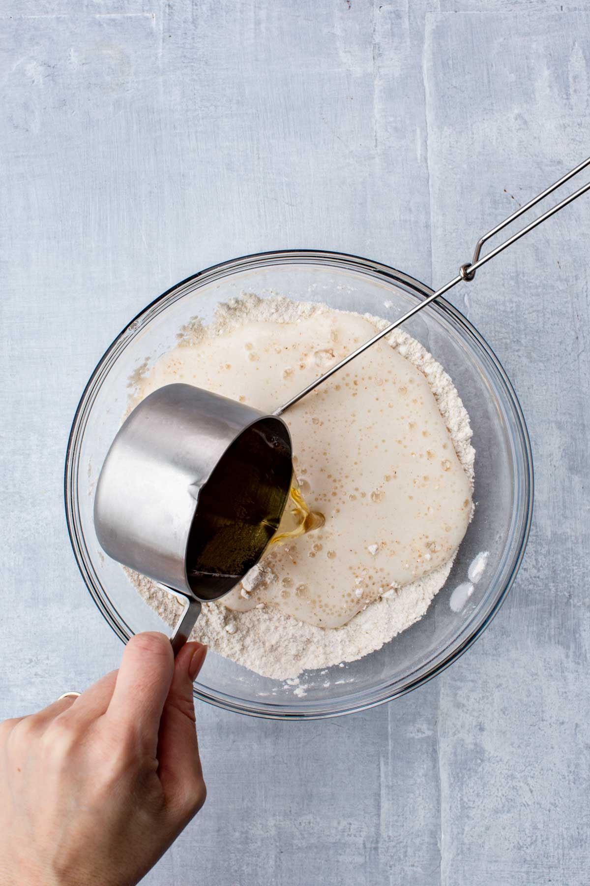 Hand pouring beer and liquid into flour mixture from a metal measuring cup, starting the batter-making process.