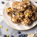 A striped plate full of oatmeal cups with blueberries and almonds, surrounded by oats, fresh blueberries, and yellow flowers.