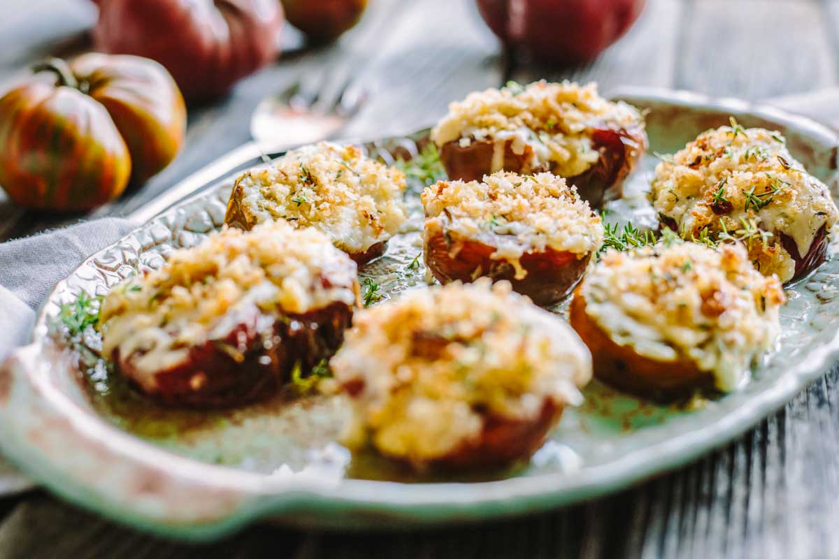 Halved heirloom tomatoes baked with melted cheese and golden panko crumbs, served on a rustic ceramic platter with fresh herbs.